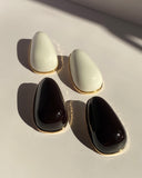 Black sculptural teardrop stud earrings with polished gold-tone border displayed on clear jewelry stand