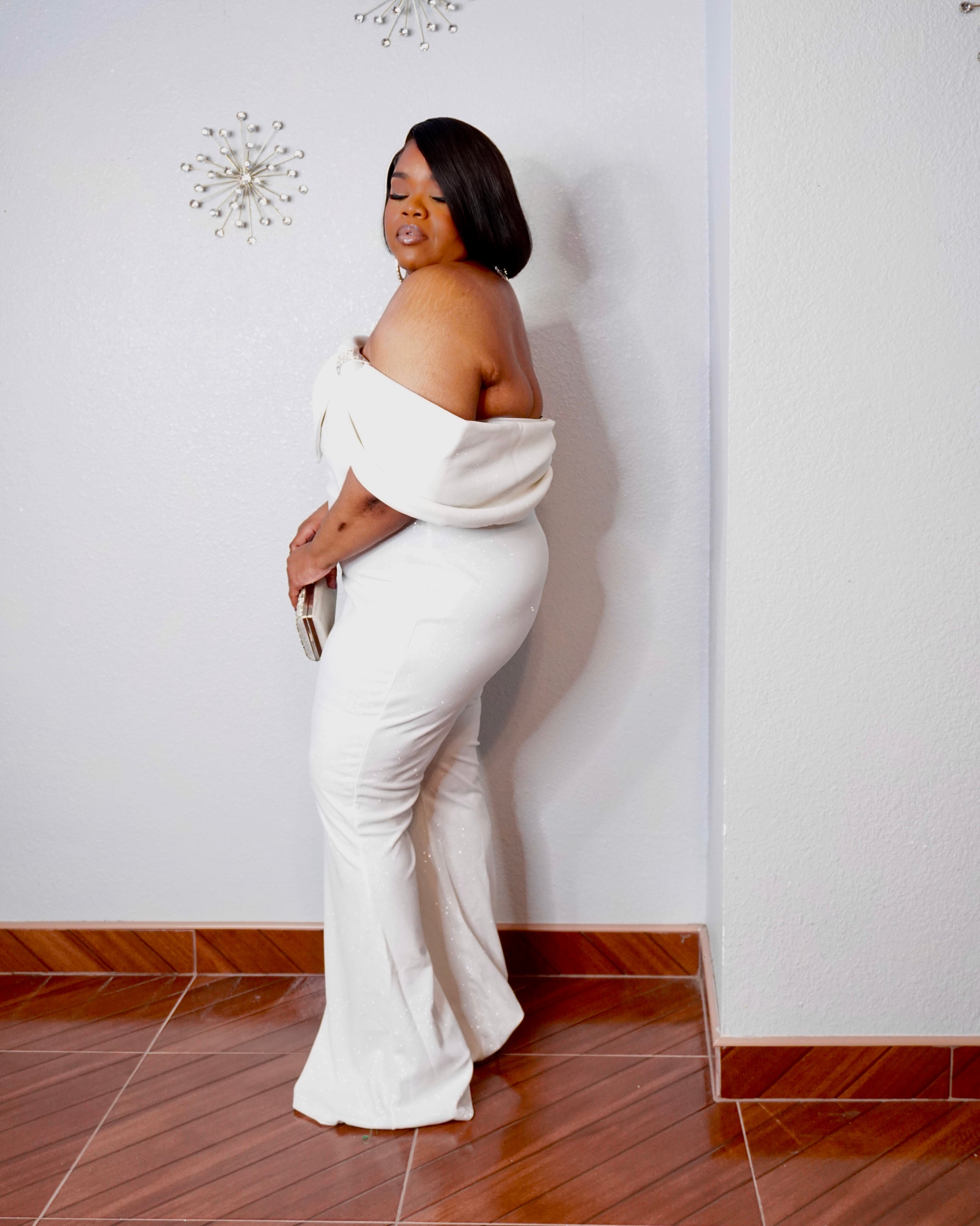 Plus size model wearing the LYSAIRE Ivory Empress off-shoulder formal jumpsuit in ivory white, displaying the dramatic draped sleeve, sparkling shoulder embellishment, asymmetrical neckline, tailored fitted bodice, and elegant full-length wide-leg trousers, paired with a silver embellished clutch for a sophisticated wedding guest or formal evening look.
