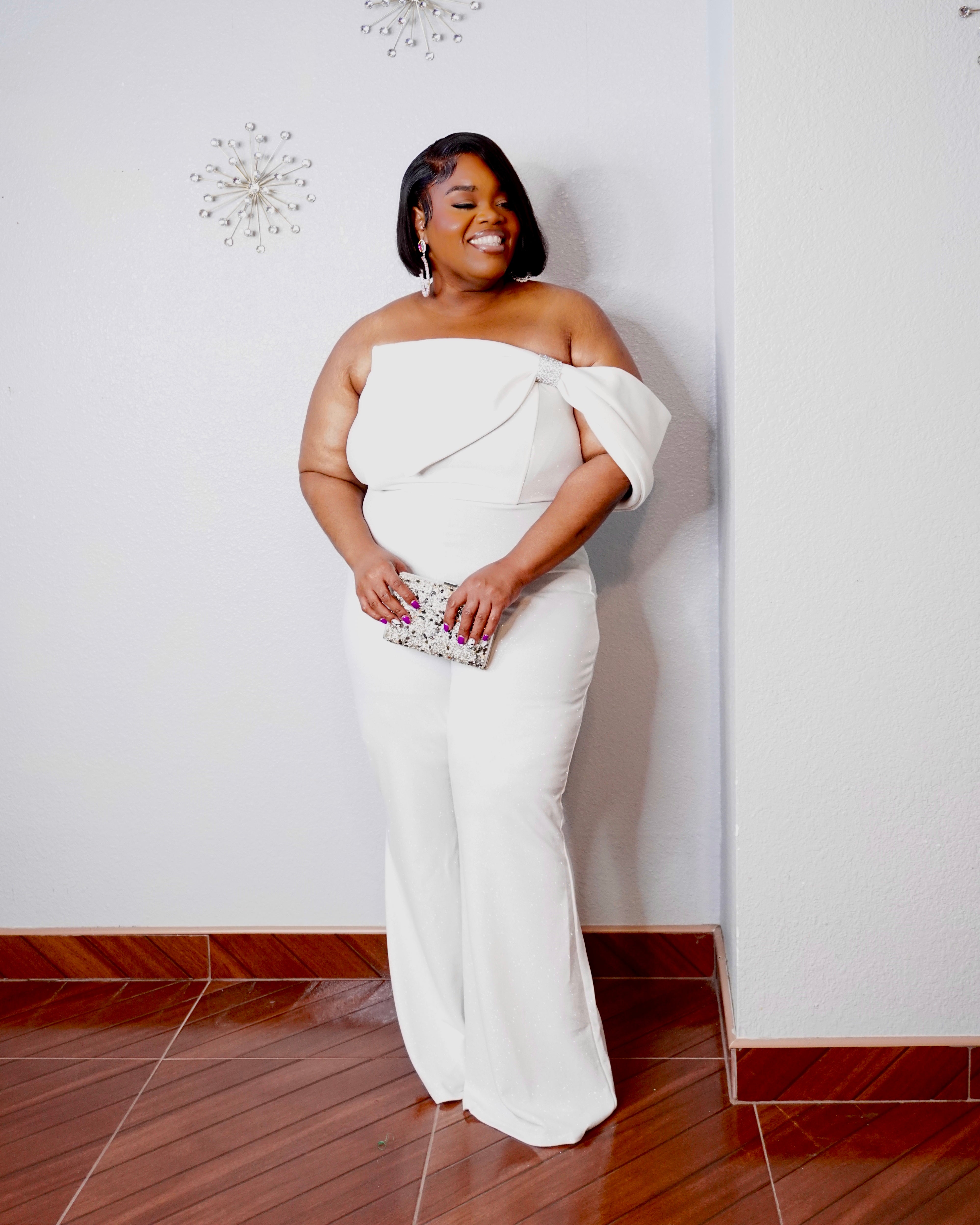Side-angle full-length view of the LYSAIRE Ivory Empress plus size ivory-white formal jumpsuit, featuring a sculpted strapless neckline, elegant asymmetrical bodice draping, flattering fitted waist and hips, and sophisticated wide-leg pants, styled with sparkling statement earrings, metallic heels, and a crystal clutch for a glamorous evening ensemble.