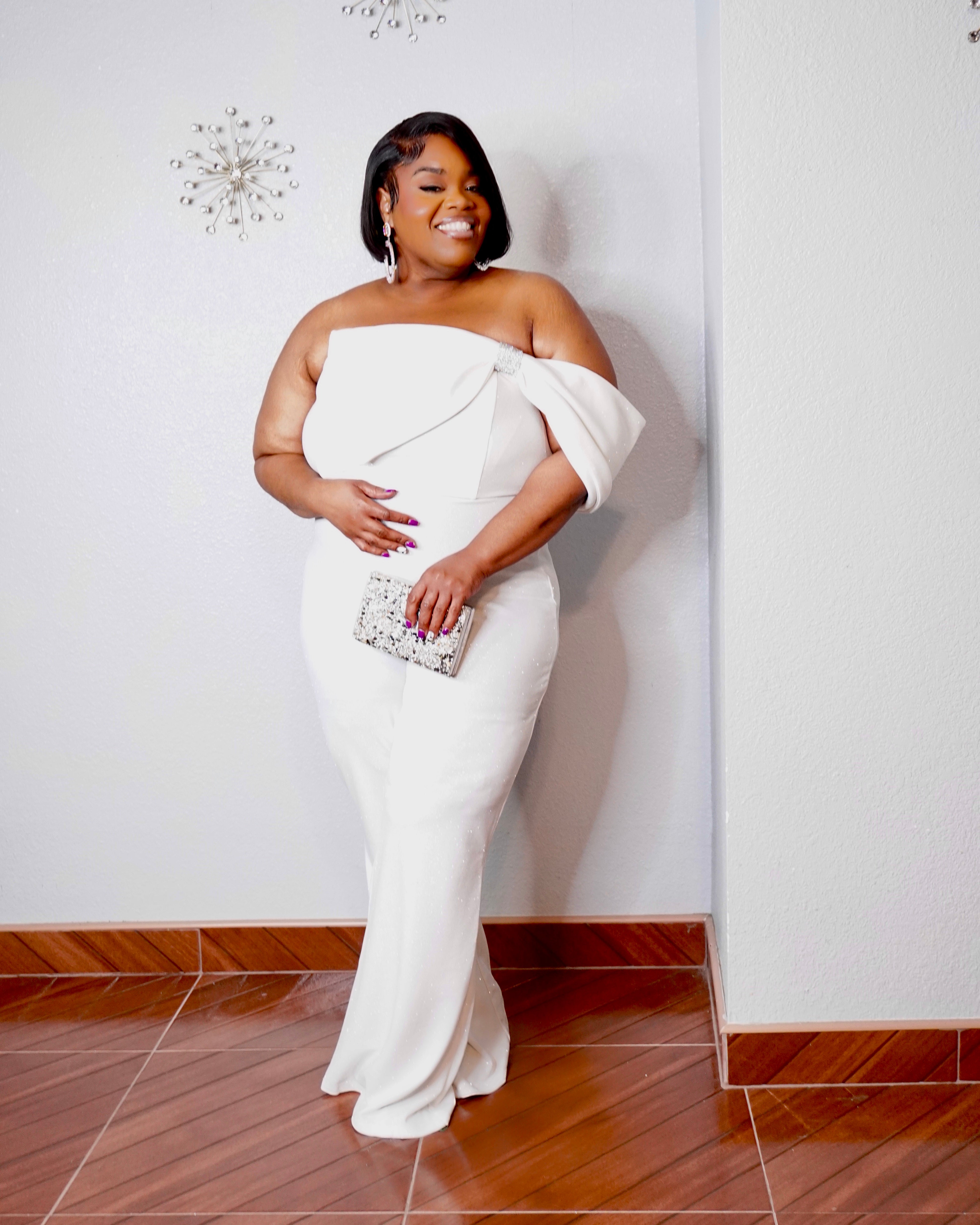 Rear three-quarter view of the LYSAIRE Ivory Empress plus size off-shoulder jumpsuit, showcasing the softly draped sleeve wrapping around the upper arms, elegant exposed back neckline, fitted waist and hip silhouette, subtle fabric shimmer, and long flowing wide-leg pants, creating a sophisticated formalwear look for special occasions.
