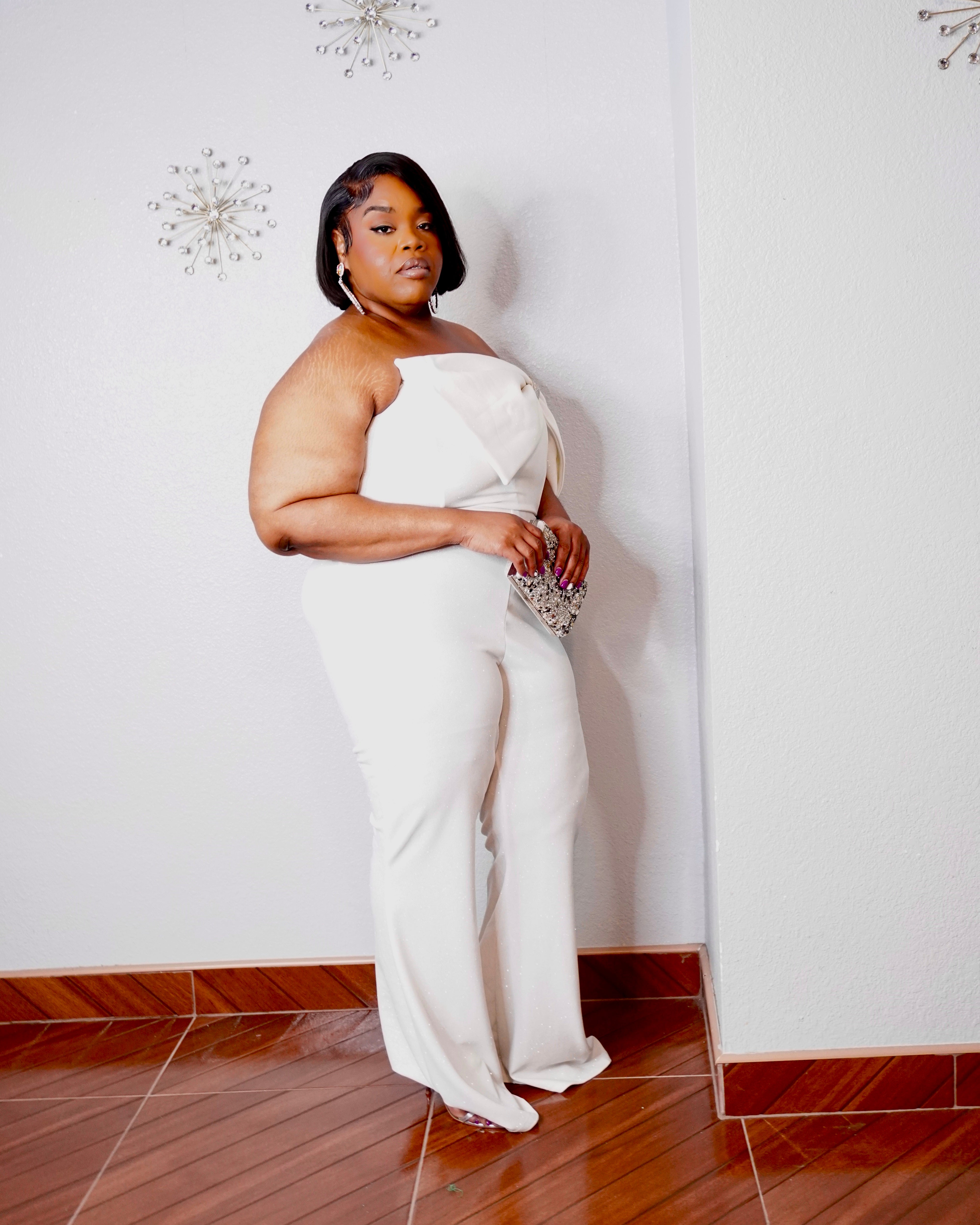 Three-quarter rear view of a plus size model wearing the LYSAIRE Ivory Empress ivory-white formal jumpsuit, highlighting the elegant off-shoulder draping across the upper arms, strapless back neckline, fitted waist, flattering hip silhouette, and full-length wide-leg pants crafted from subtly shimmering fabric.