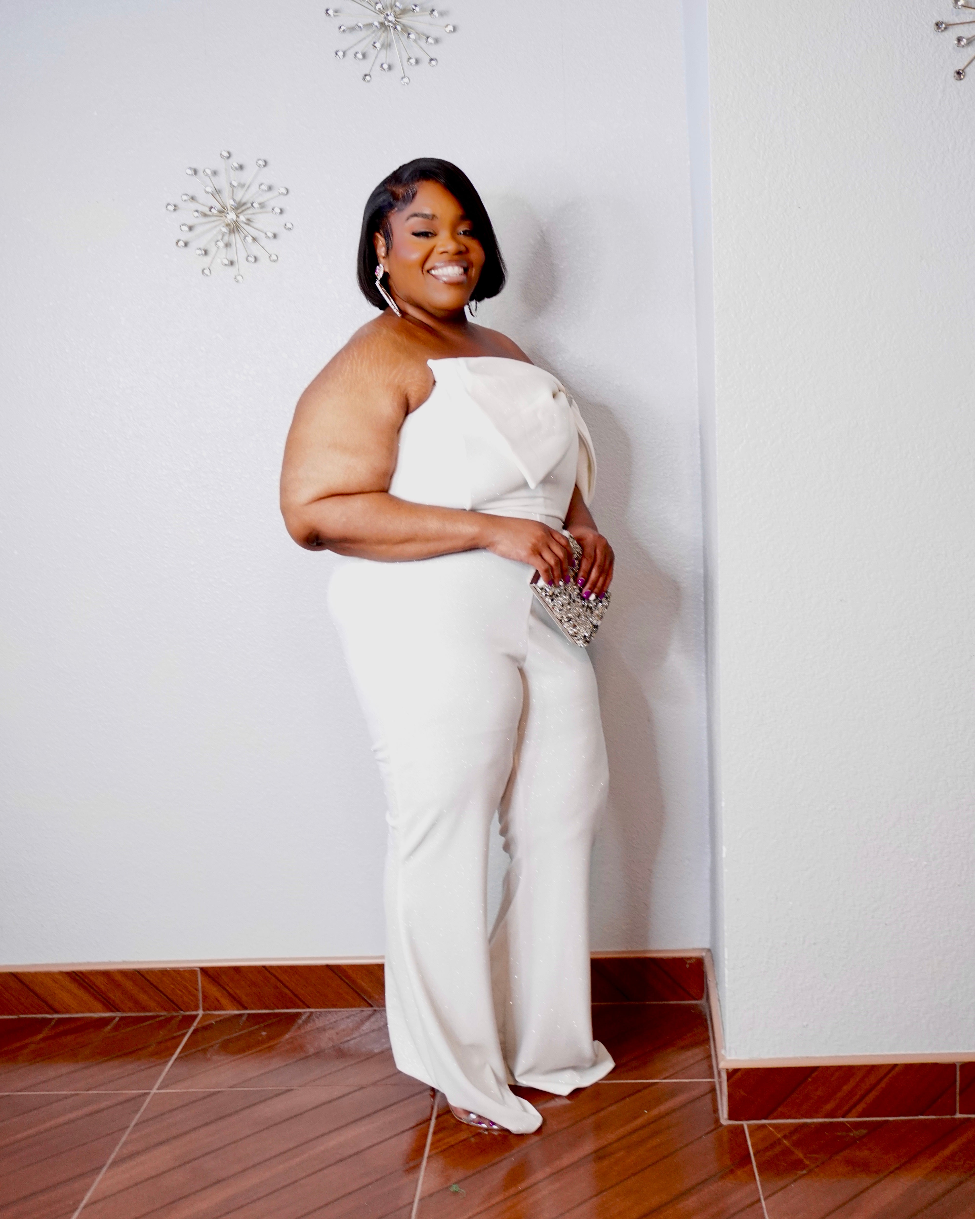 Full-length back view of the LYSAIRE Ivory Empress plus size formal jumpsuit in ivory white, displaying the strapless back design, center-back zipper closure, dramatic draped off-shoulder sleeve, fitted waist and hips, and flowing wide-leg trousers, offering a complete look at the elegant construction and silhouette of this statement evening outfit.