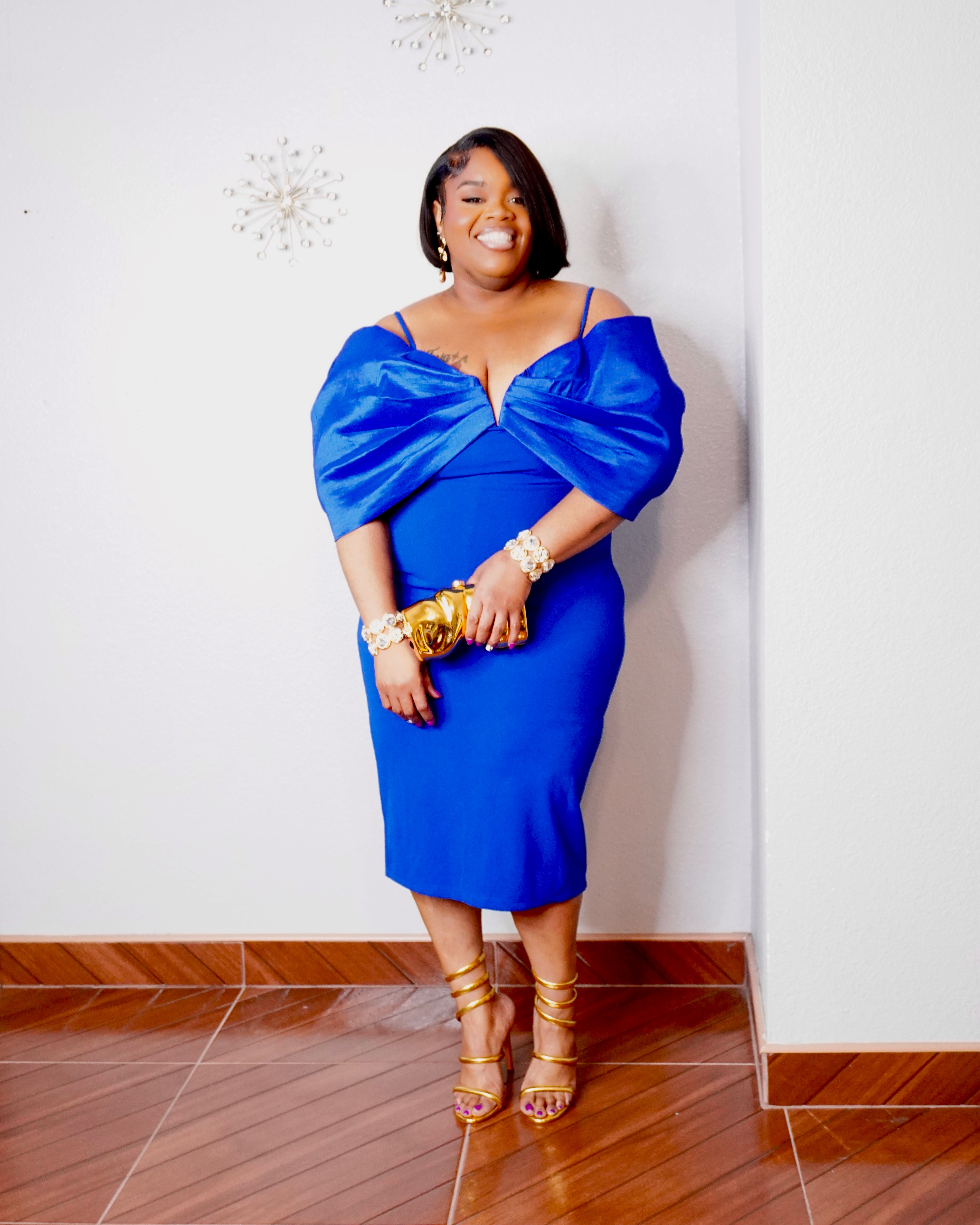 Plus size model wearing a royal blue fitted cocktail dress with oversized draped sleeves, gold statement jewelry and strappy heels.