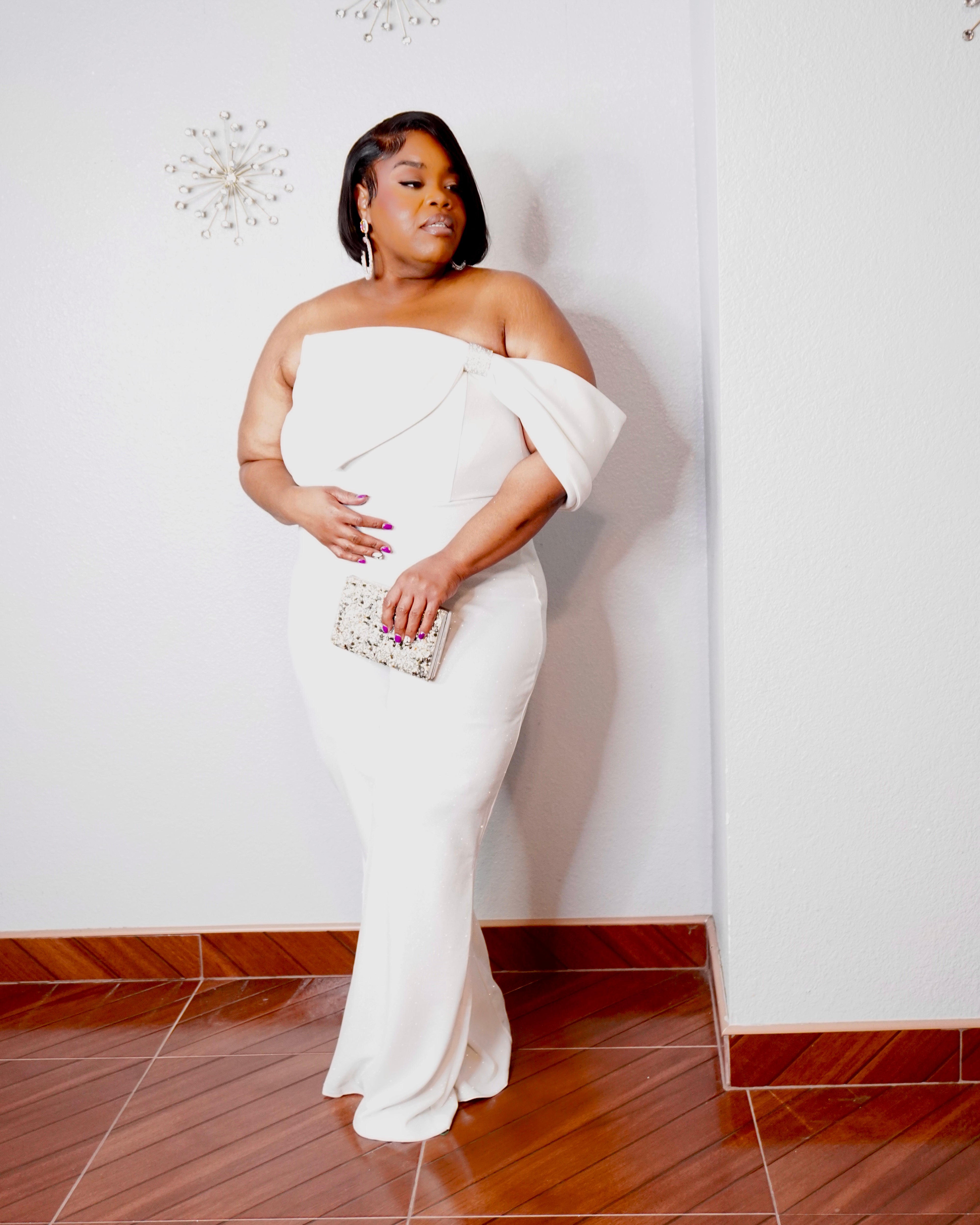 Side-profile view of the LYSAIRE Ivory Empress plus size white formal jumpsuit, showcasing the dramatic off-shoulder draped fabric, exposed shoulder neckline, fitted waist and hip silhouette, subtly shimmering material, and flowing wide-leg pants, creating an elegant and sophisticated outfit for formal occasions, gala events, and evening celebrations.